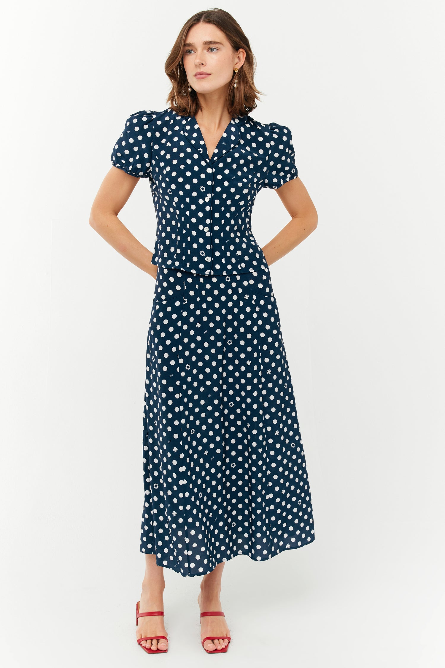 A model wears a two piece set with a collared short sleeved shirt and a matching high waisted long skirt. The print is white polkadots on a dark navy base. She wears red shoes.