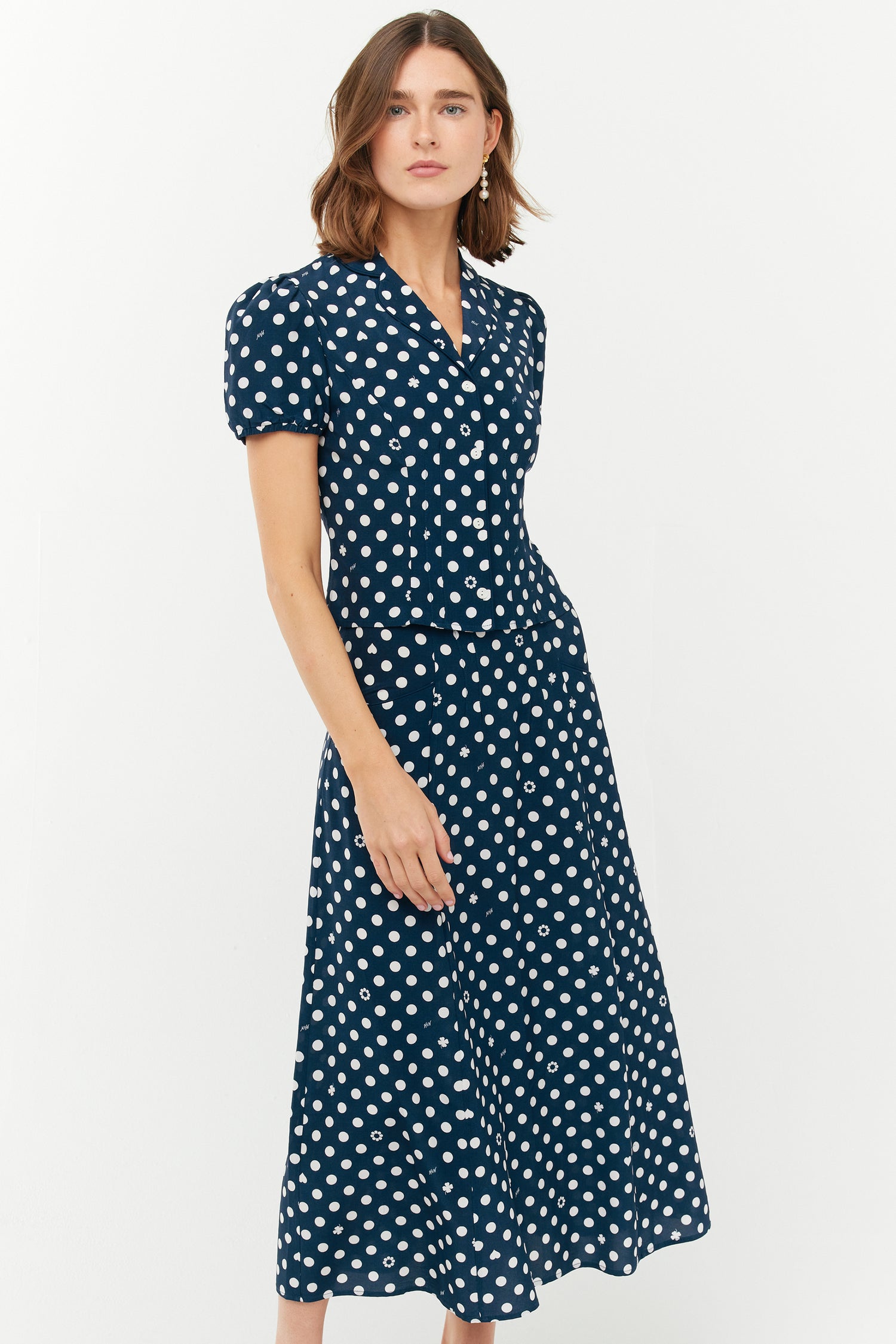 A model wears a two piece set with a collared short sleeved shirt and a matching high waisted long skirt. The print is white polkadots on a dark navy base.