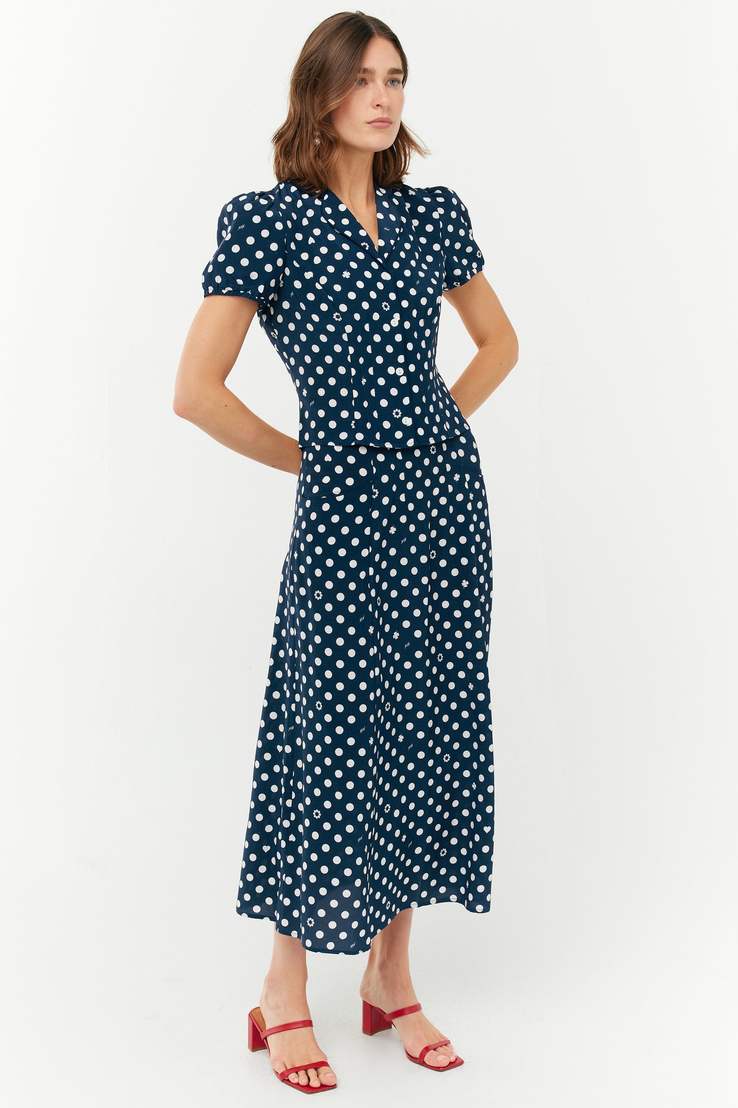 A model wears a two piece set with a collared short sleeved shirt and a matching high waisted long skirt. The print is white polkadots on a dark navy base. She wears red shoes.