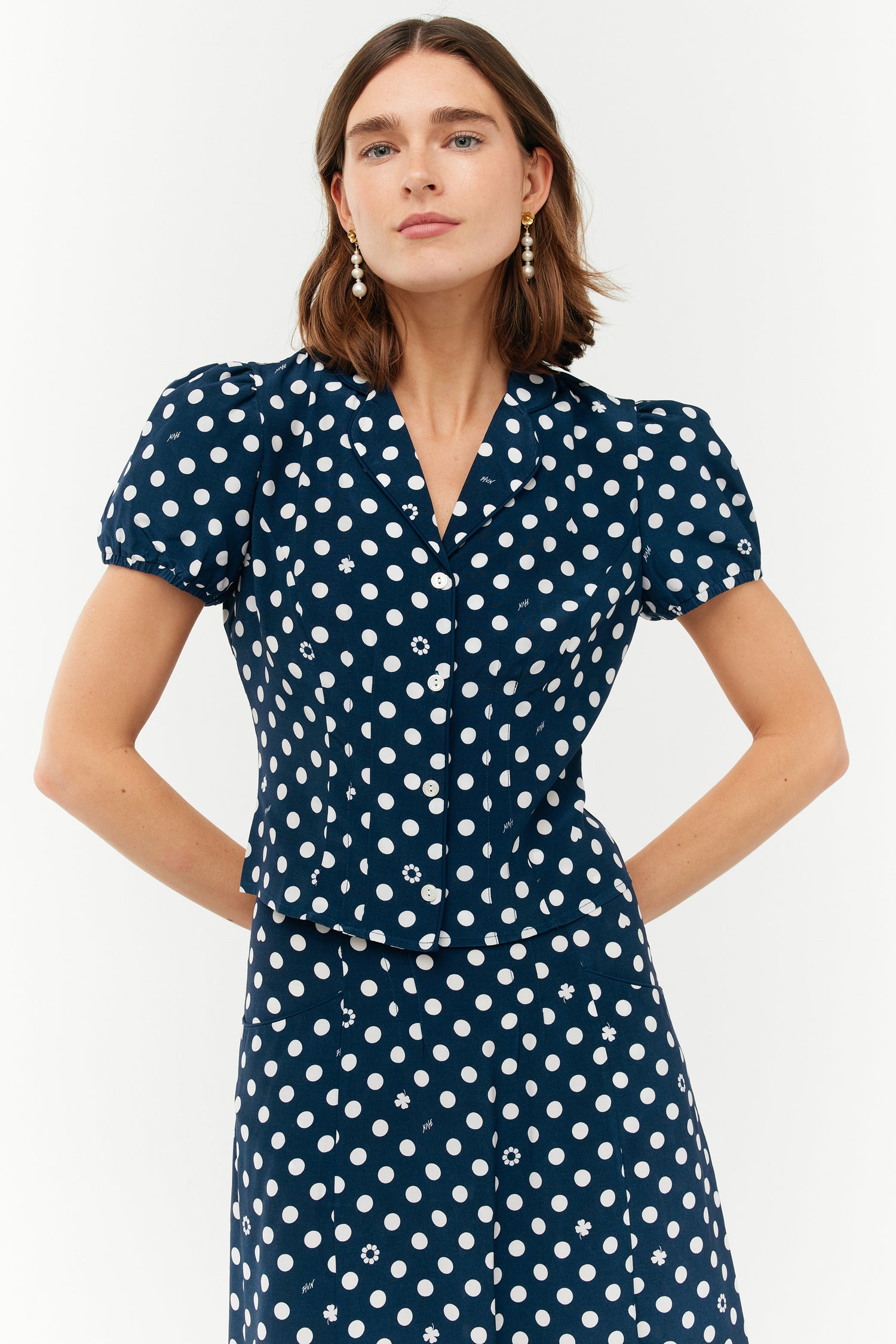 A model wears a two piece set with a collared short sleeved shirt and a matching high waisted long skirt. The print is white polkadots on a dark navy base.