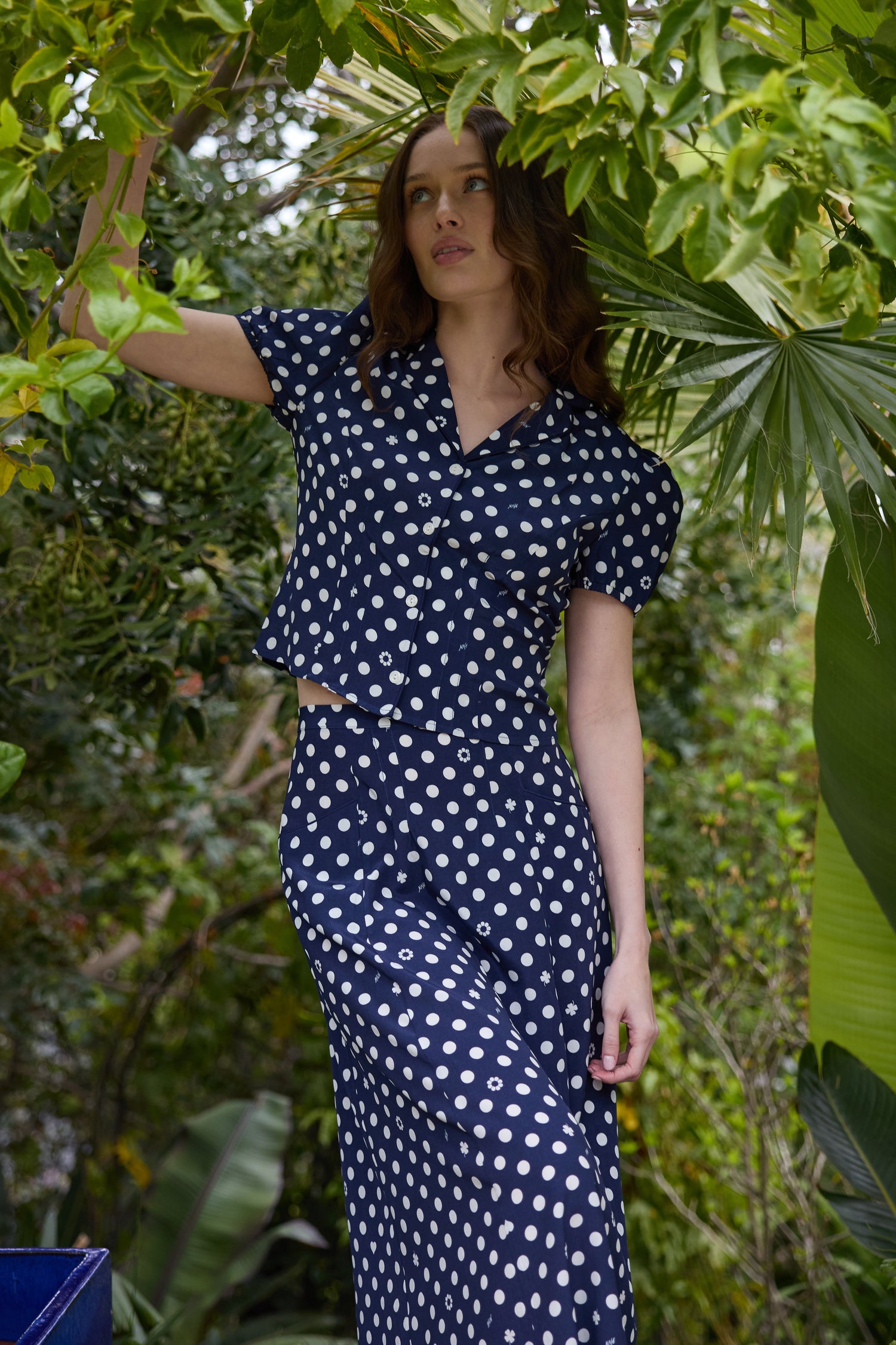 A model wears a two piece set with a collared short sleeved shirt and a matching high waisted long skirt. The print is white polkadots on a dark navy base. She is in a garden.