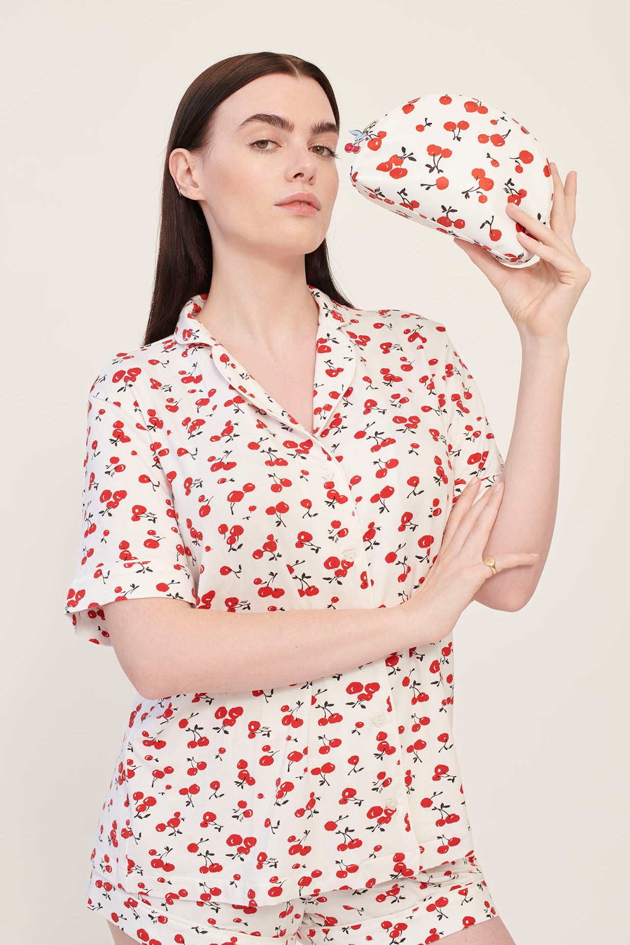 A model wearing cherry print pajamas holds a cherry print cosmetic case with a cherry zip pull.