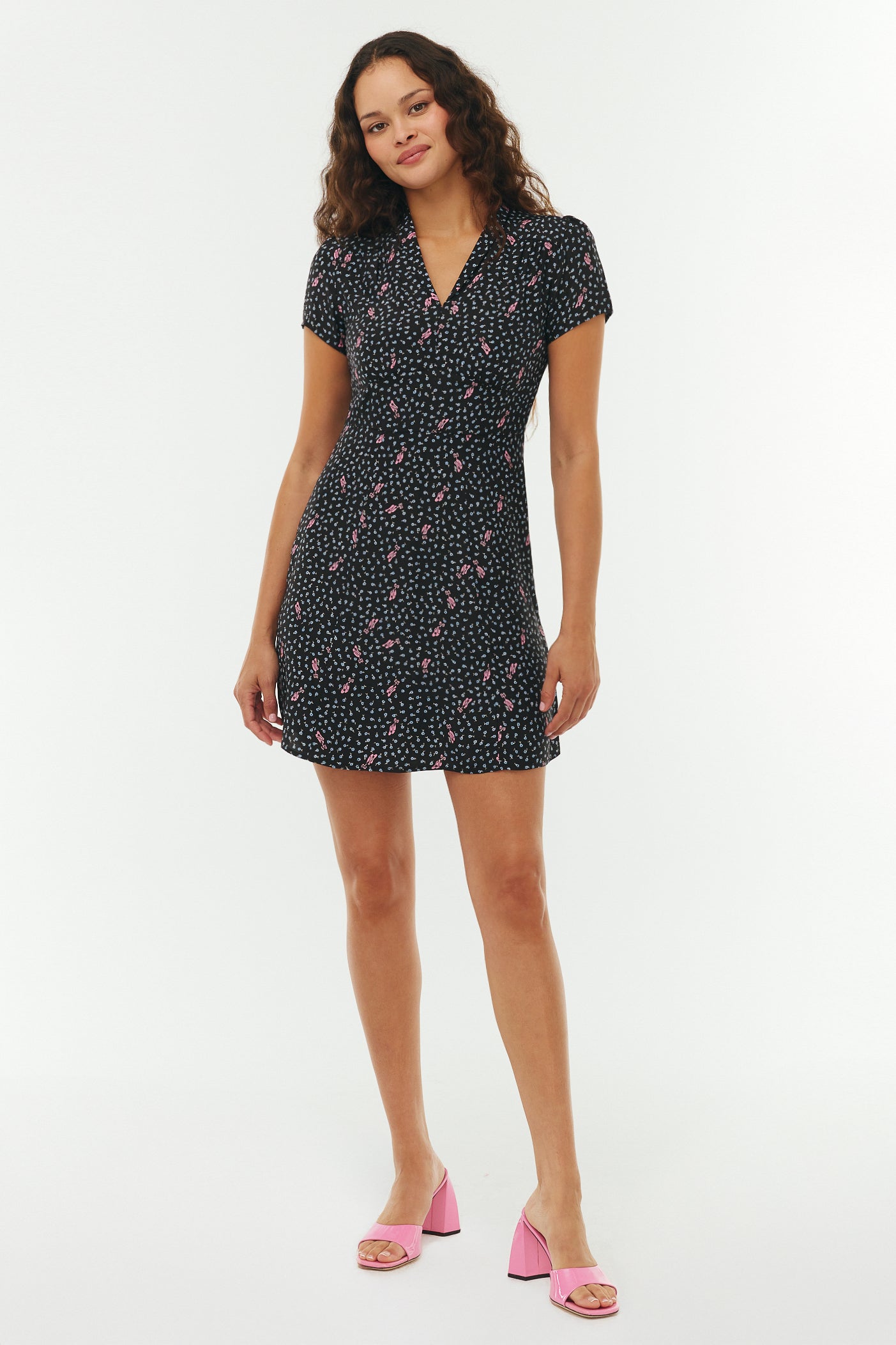 Woman wearing a black floral mini dress with pink ballet slippers on a white background. She wears pink heels. 