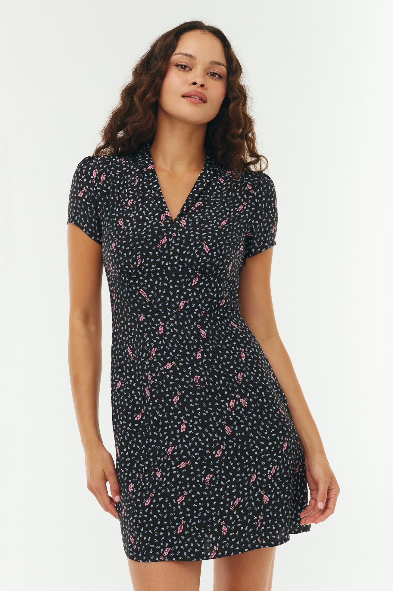 Woman wearing a black floral mini dress with pink ballet slippers on a white background. 