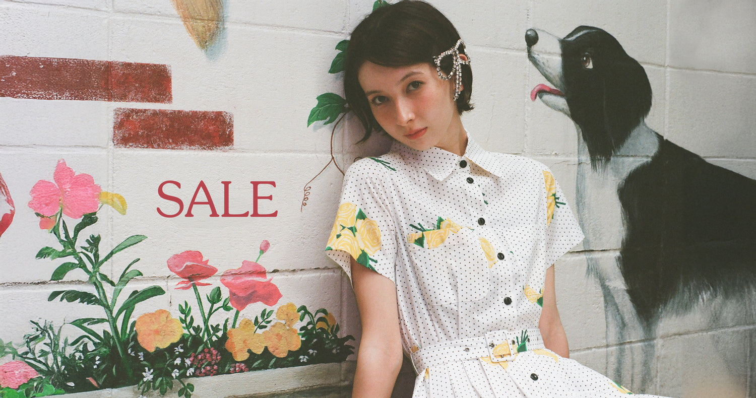 A girl in a collared button up dress with a belt sits on the floor. She sits in front of a painted mural with a dog with his tounge out and flowers. It says SALE in big letters by her face. 