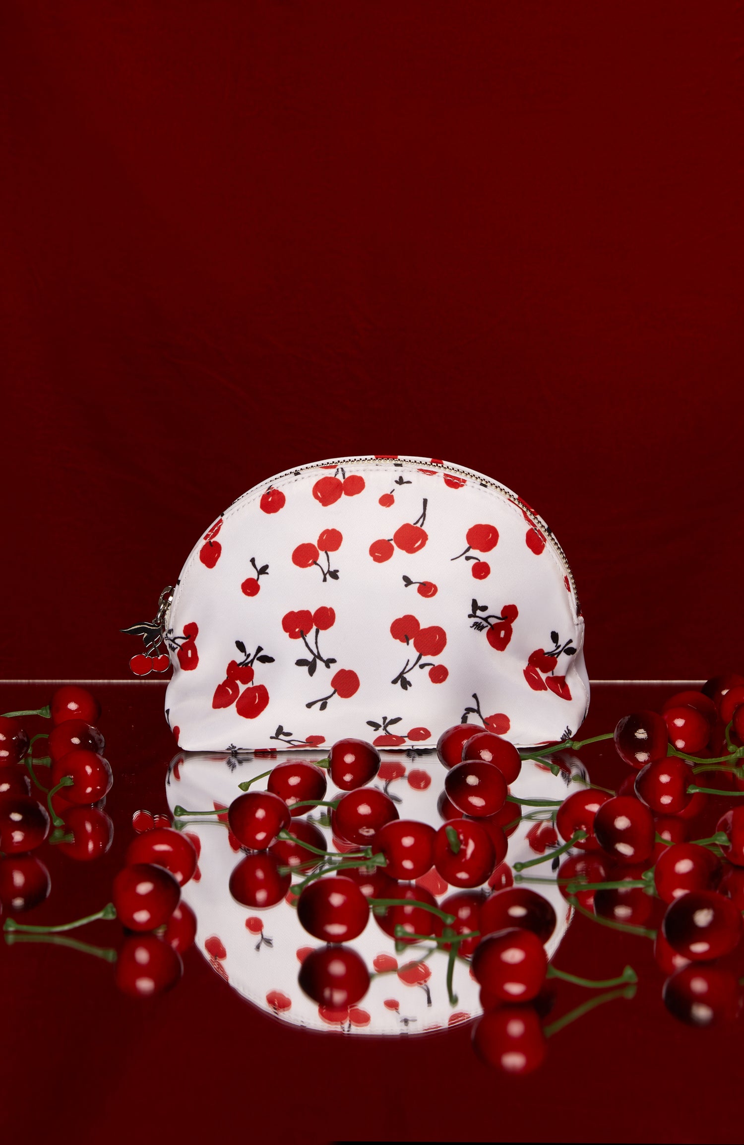 Acherry print cosmetic case with a cherry zip pull surrounded by cherries on a red background.