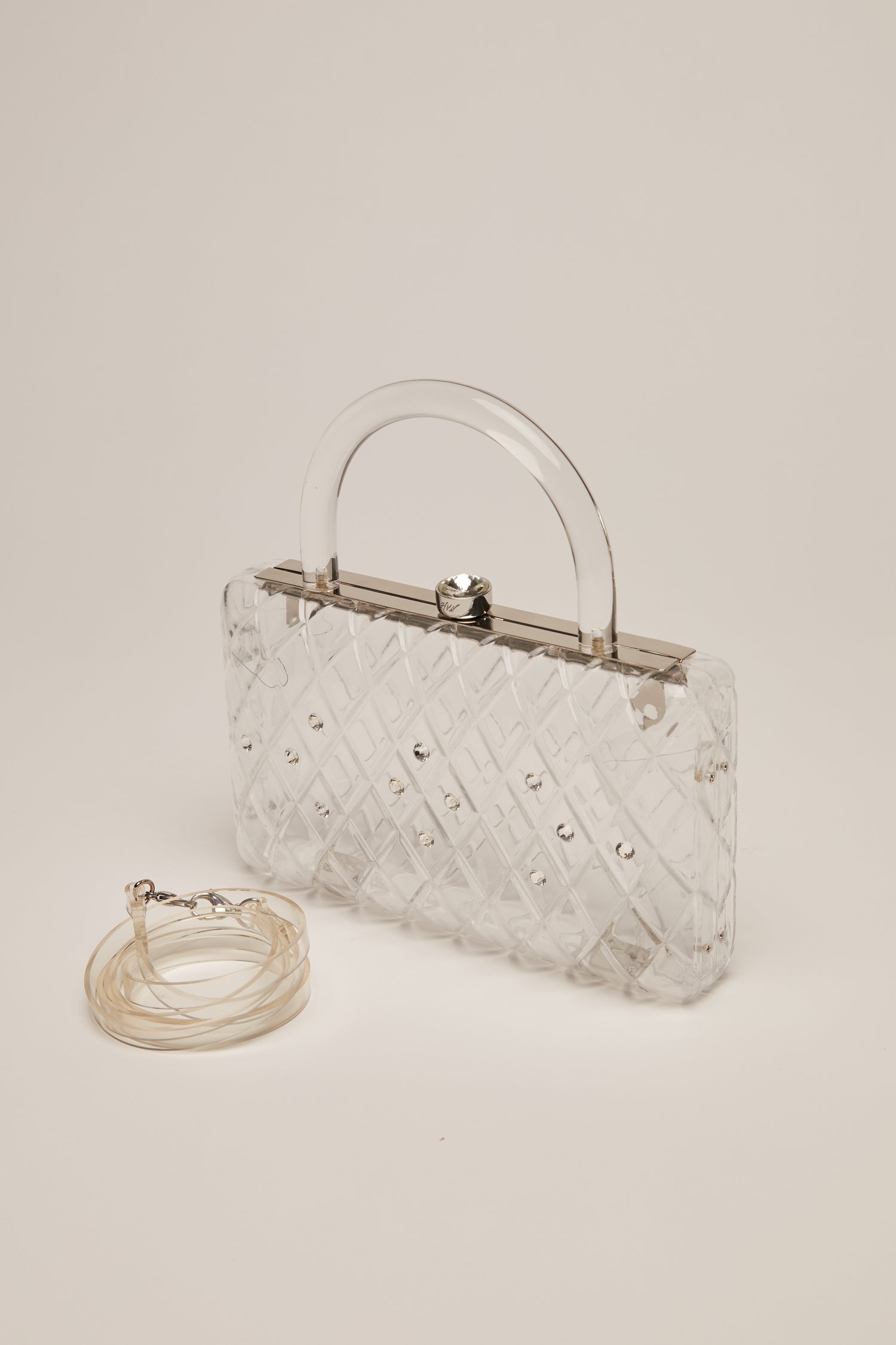 A clear acrylic handbag with crystals and a short handle with a curled up strap next to it. 
