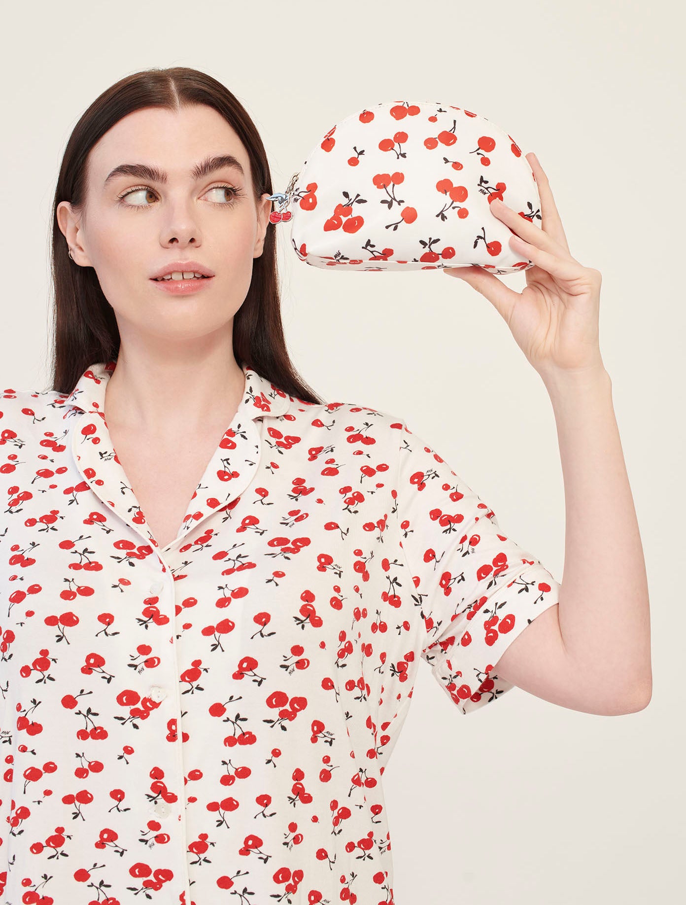 A model wearing cherry print pajamas holds a cherry print cosmetic case with a cherry zip pull. 
